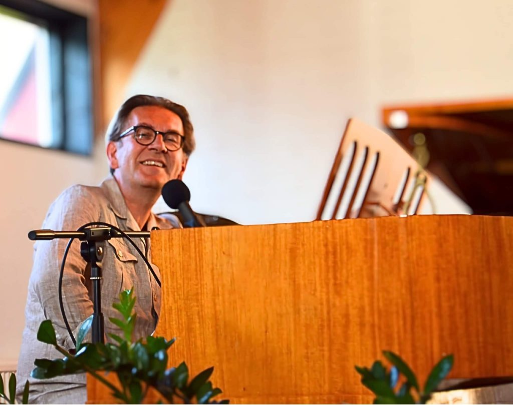 En man ler medan han spelar på en orgel i en väl upplyst inomhusmiljö med gröna växter i förgrunden.