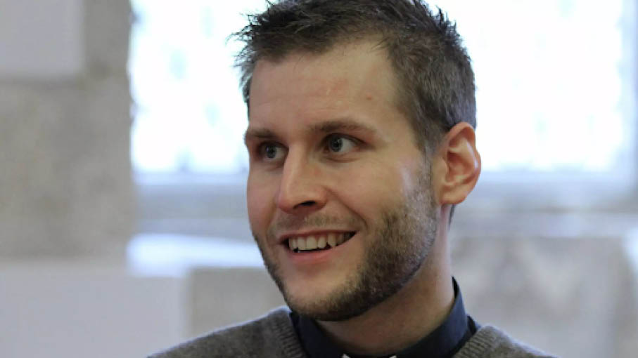 En man med kort hår och skägg ler, klädd i skjorta med krage och tröja, inomhus med dämpad belysning.