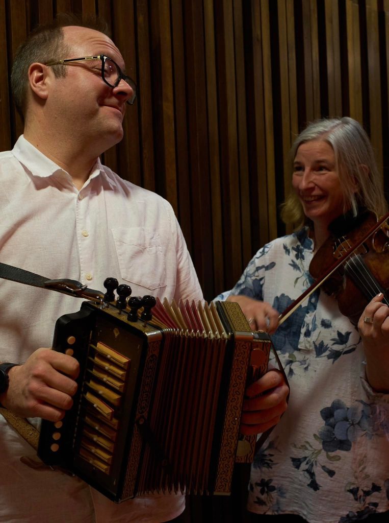 En man som spelar dragspel och en kvinna som spelar violin ler mot varandra mot en trävägg.