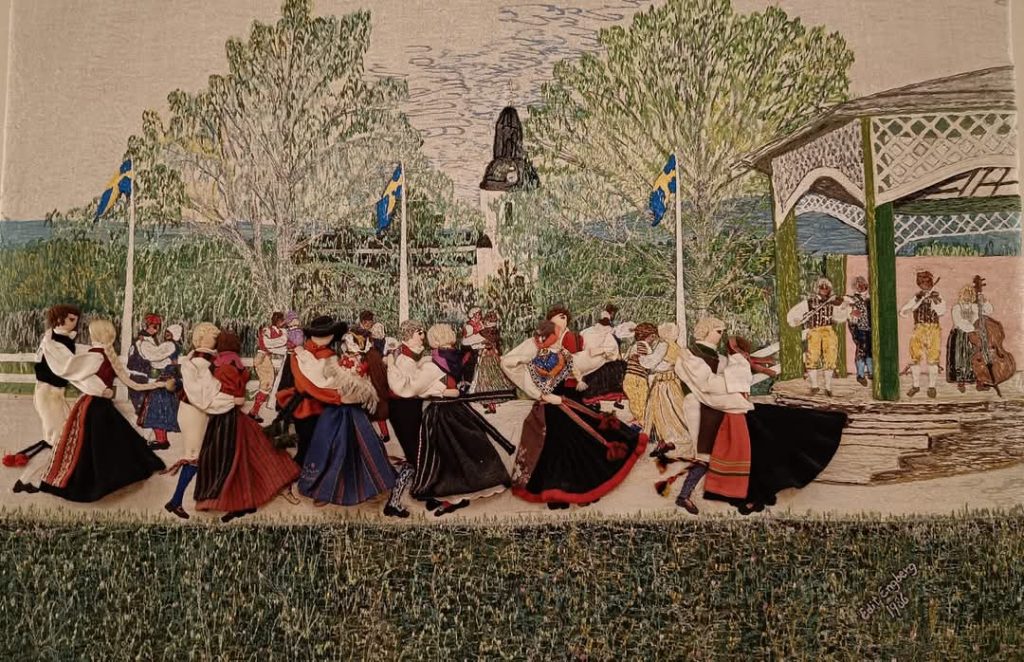 Människor i traditionella svenska kläder dansar utomhus nära musiker och svenska flaggor en solig dag.