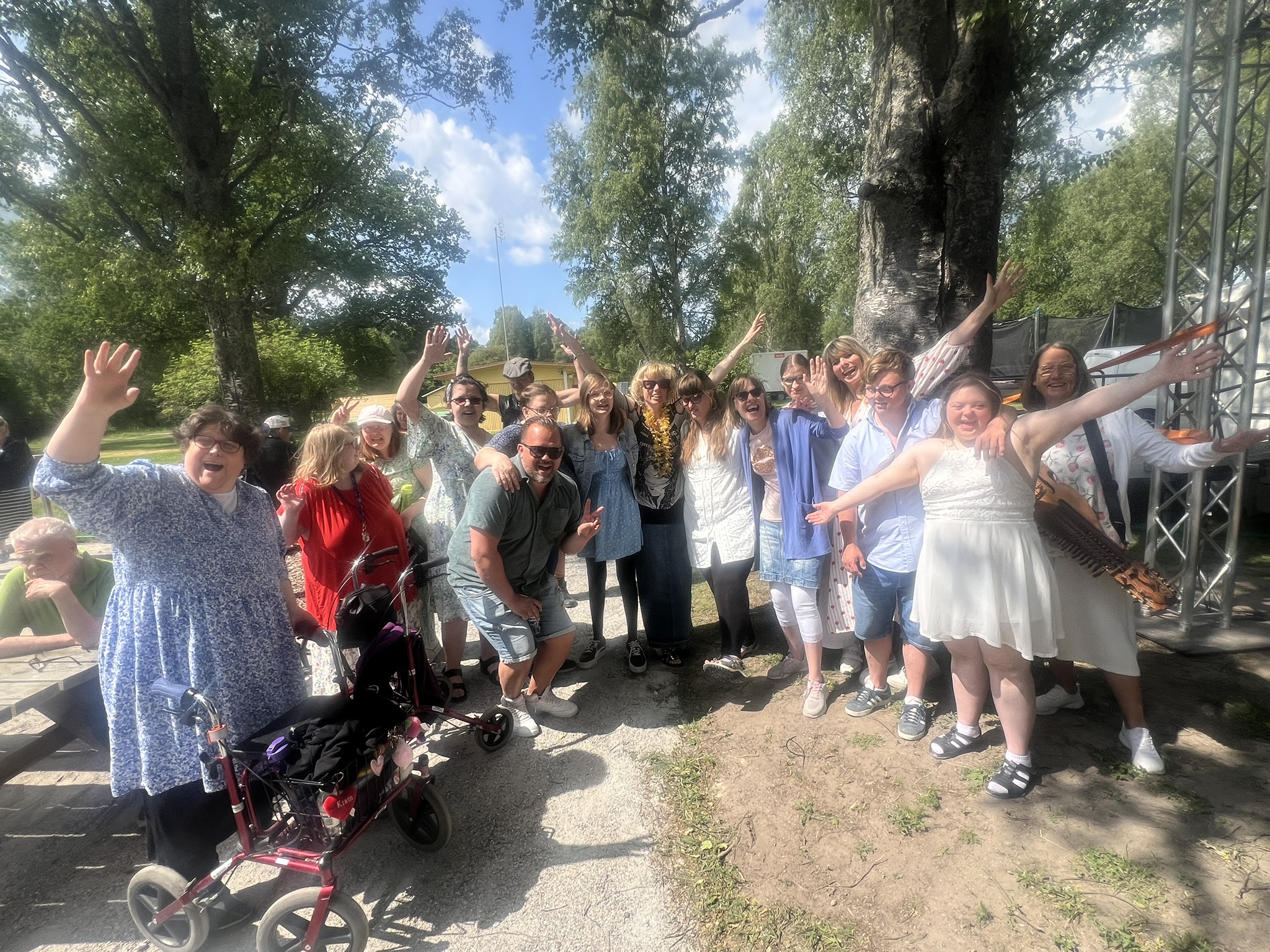 En grupp människor utomhus ler och vinkar och poserar tillsammans en solig dag under höga träd.