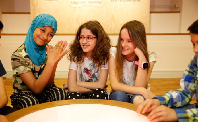 Fyra leende barn sitter tillsammans runt ett runt bord och pratar och njuter av varandras sällskap.