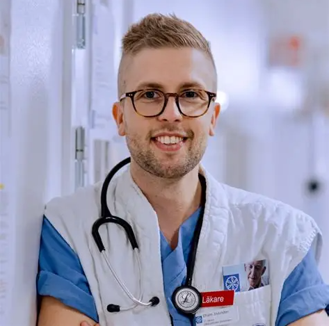 En leende läkare med glasögon och stetoskop står i en sjukhuskorridor.