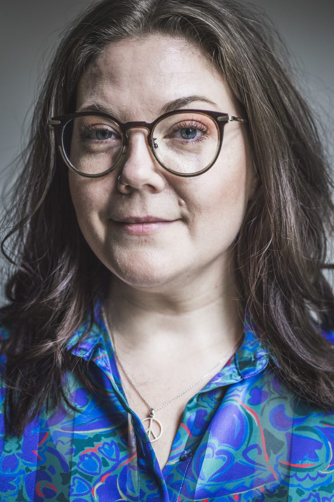 Kvinna med långt brunt hår, glasögon och blåmönstrad skjorta, ler lätt mot kameran.