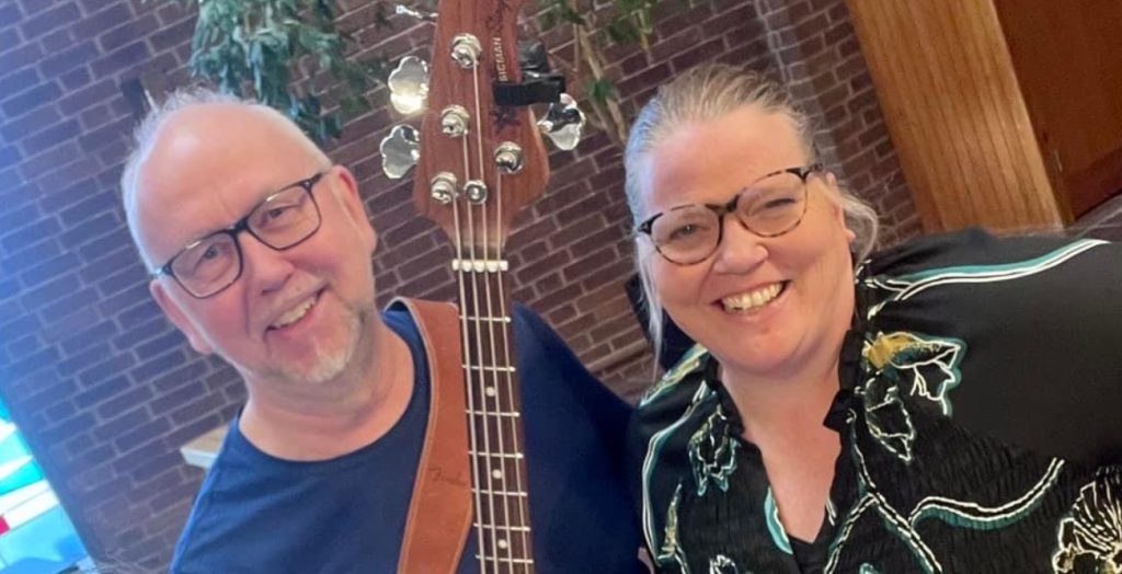 Två leende personer med glasögon poserar inomhus, den ena håller i en gitarr. Tegelvägg och växt i bakgrunden.