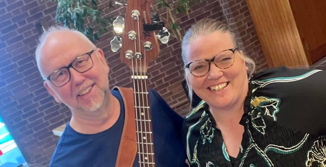 Två leende personer med glasögon poserar inomhus, den ena håller i en gitarr. Tegelvägg och växt i bakgrunden.