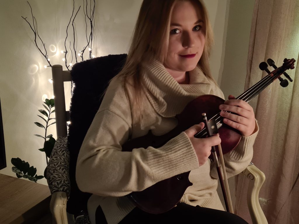 Kvinna sitter på en stol och håller i en fiol, med ljusslingor och växter i bakgrunden.