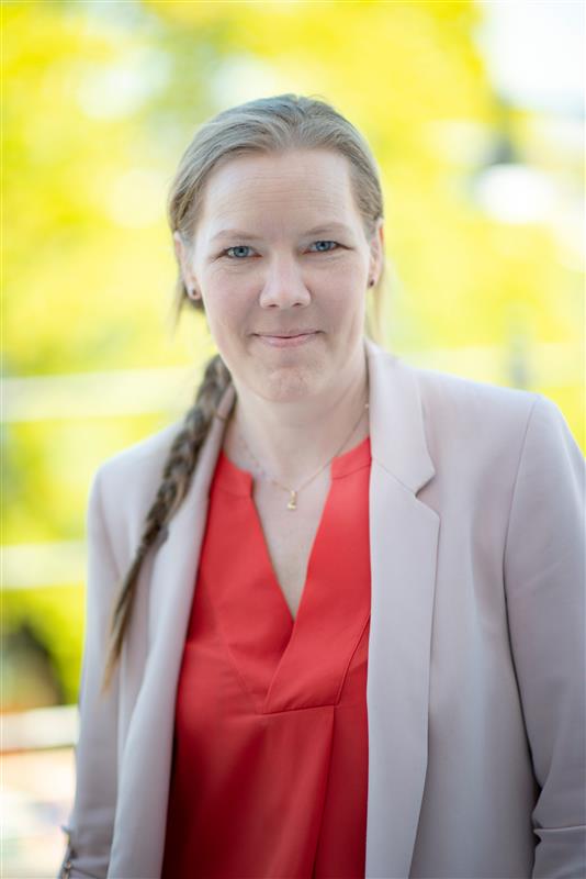 Kvinna med lång fläta i röd blus och ljus kavaj, står utomhus mot en suddig bakgrund.