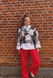 Person i röda byxor, vit skjorta, mönstrad jacka och glasögon står leende mot en tegelvägg.