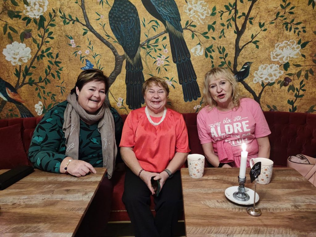 Tre kvinnor sitter vid ett bord på ett kafé, leende, med tekoppar och ett tänt ljus framför sig.