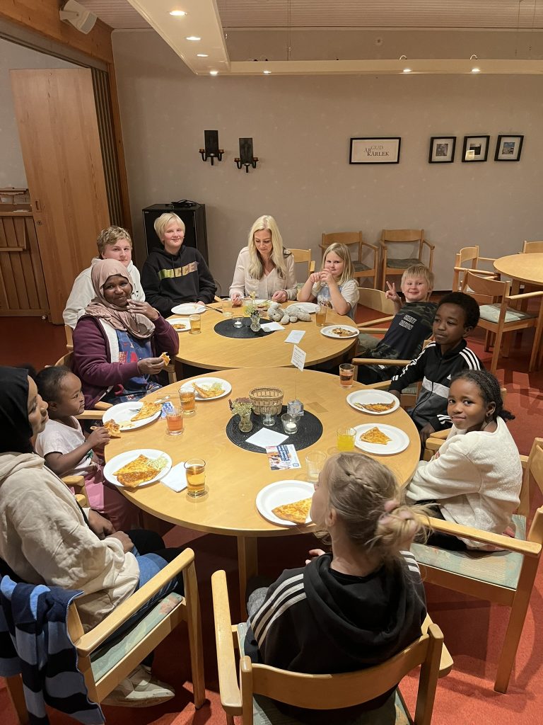 En grupp barn och en vuxen sitter runt borden, äter mat och ler i en mysig matsal.