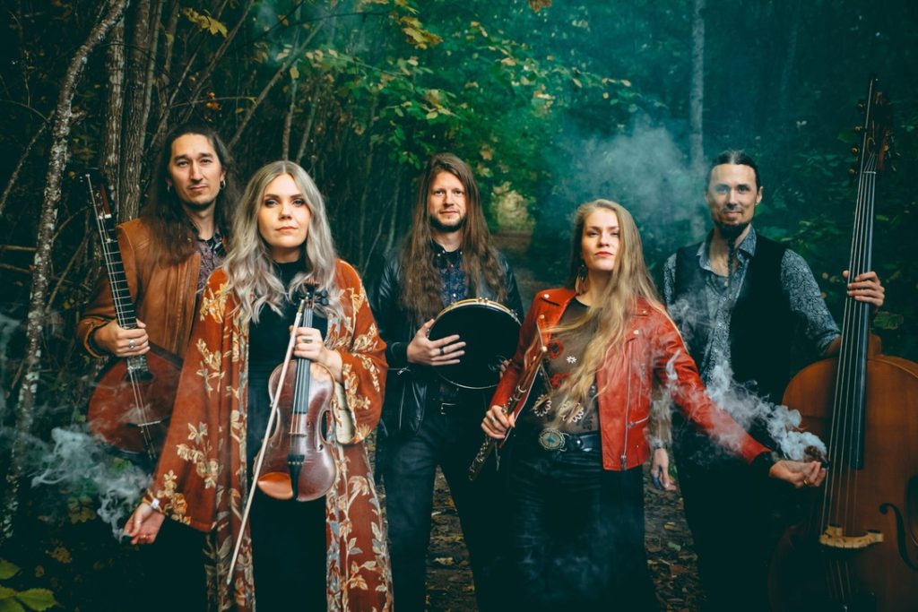 Fem musiker med långt hår står i en dimmig skog och håller i instrument som violin, tamburin och cello.