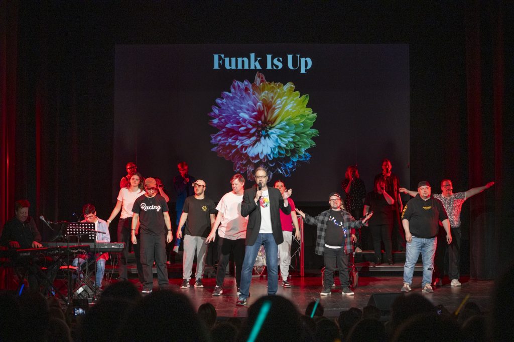 En grupp uppträder på scenen under en "Funk Is Up"-skylt med en färgglad blomstergrafik projicerad bakom dem.