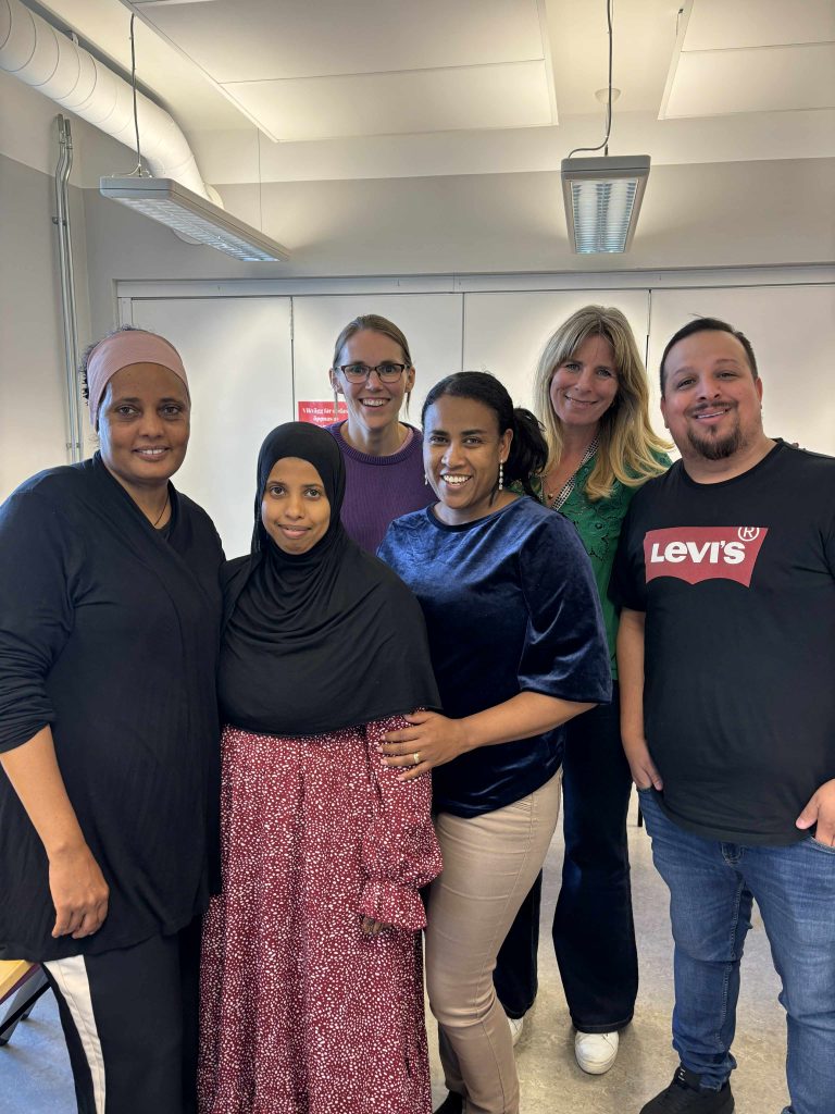 Sex vuxna poserar och ler tillsammans i ett väl upplyst klassrum med vita väggar och fluorescerande lampor.