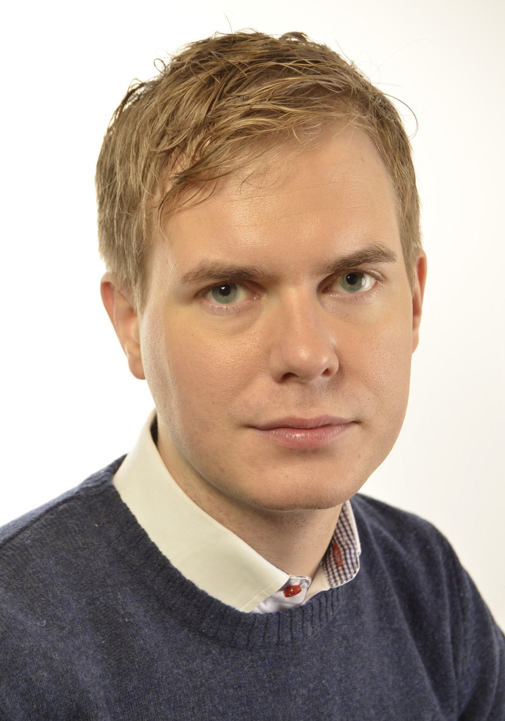 En man med kort blont hår, klädd i en blå tröja över en skjorta med krage, tittar in i kameran.