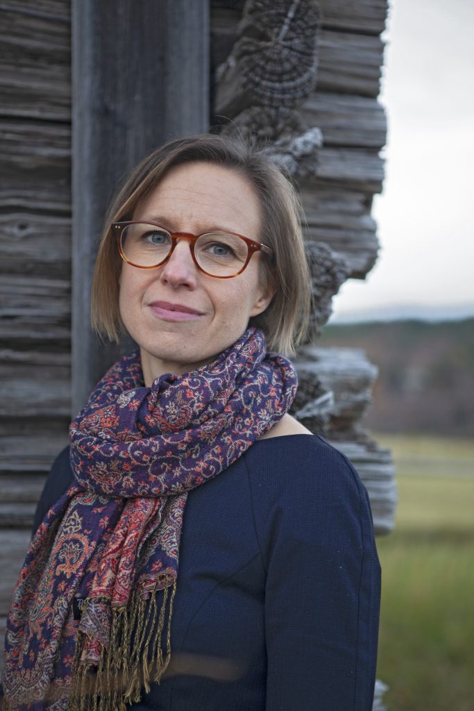 Kvinna med glasögon och mönstrad scarf står vid en trävägg och tittar lite bort från kameran.