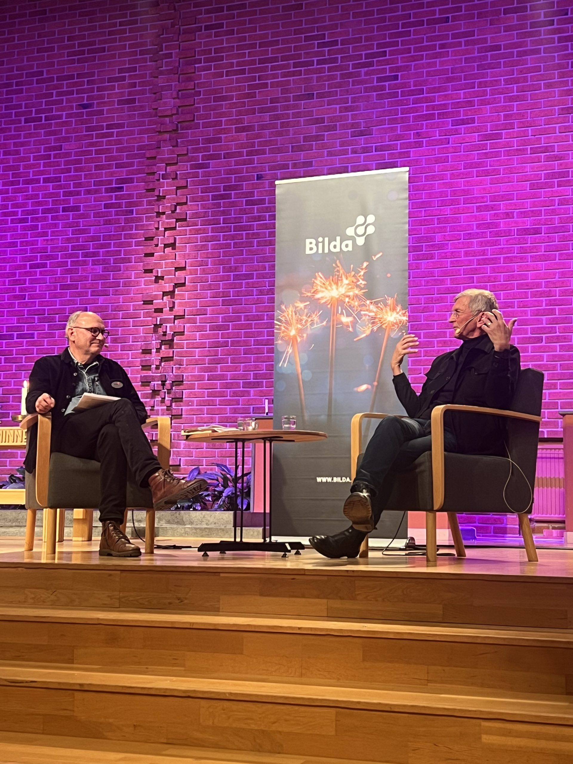 På scenen sitter två personer i fåtöljer och pratar, med en lila upplyst tegelvägg och en "Bilda"-banner bakom sig.