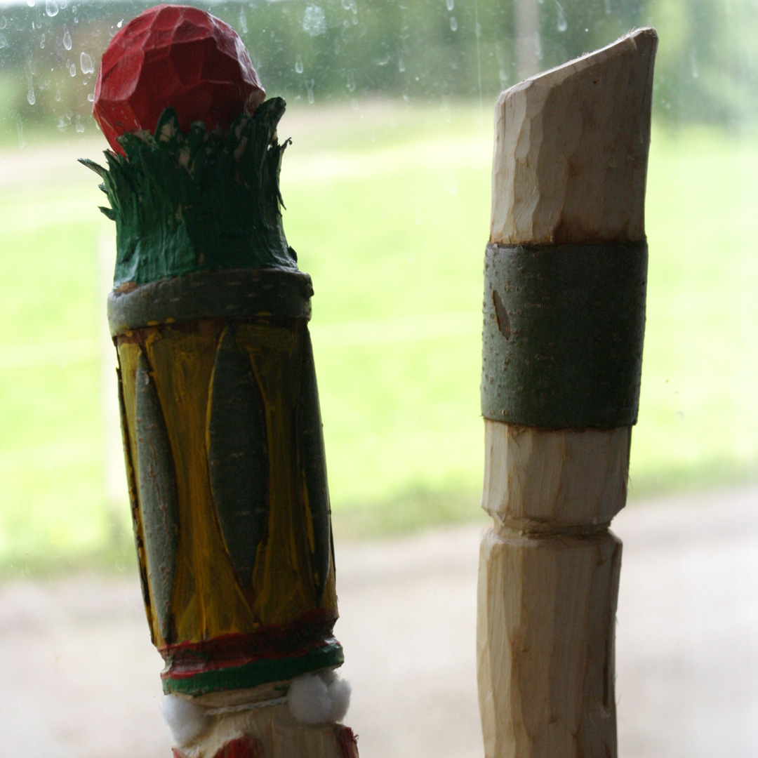 Två snidade träpinnar, den ena dekorerad med färgstark färg och en röd topp, den andra slät med ett barkband.