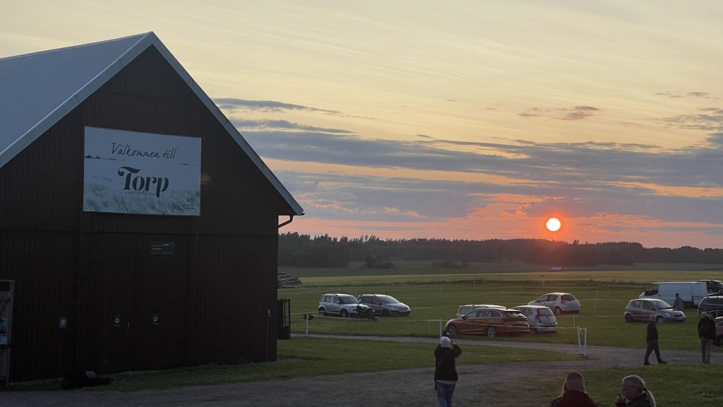 En solnedgång över en lantlig åker med parkerade bilar, människor och en lada med skylten "Välkomna till Torp".