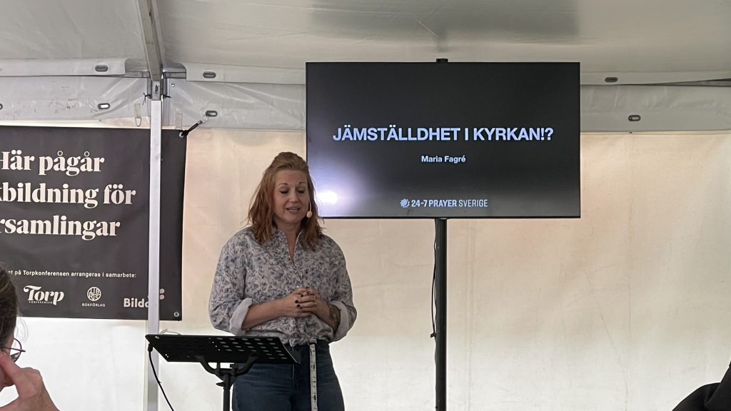 En kvinna talar på ett podium bredvid en skärm med texten "Jämställdhet i kyrkan!?" i ett vitt tält.