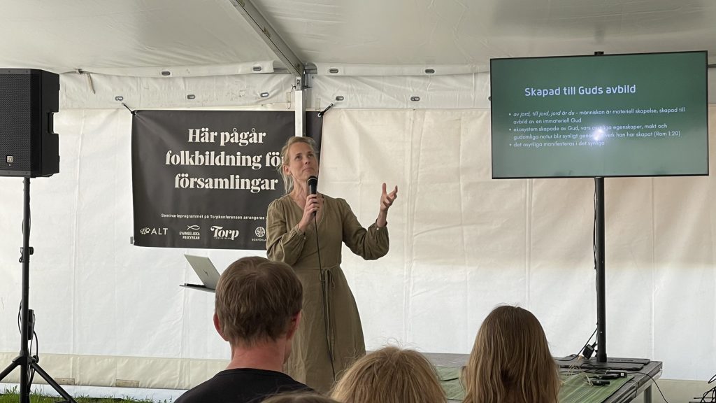 En kvinna talar på scenen med en mikrofon och gestikulerar mot en skärm, framför en publik.