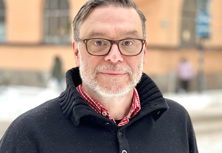 En man med glasögon och skägg, klädd i svart tröja och röd skjorta, står utomhus en snöig dag.