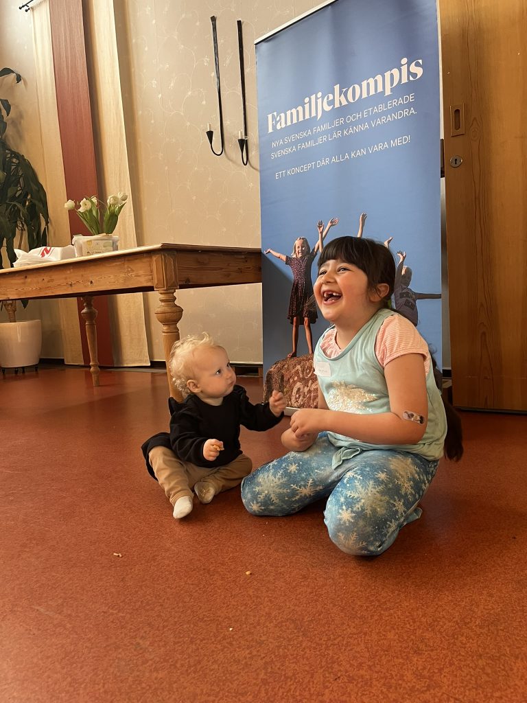 En leende flicka och en bebis sitter på golvet framför en "Familjekompis"-banderoll och håller varandra i handen.
