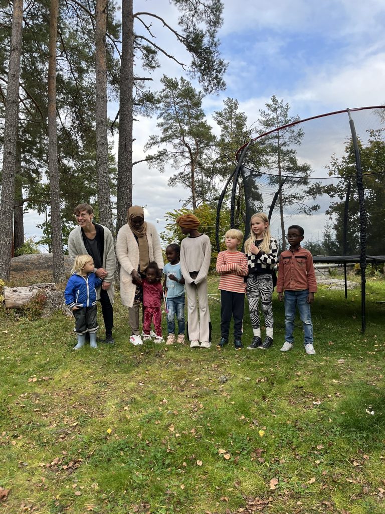 Åtta personer, både vuxna och barn, står på rad utomhus på en gräsmatta nära träd och en studsmatta.