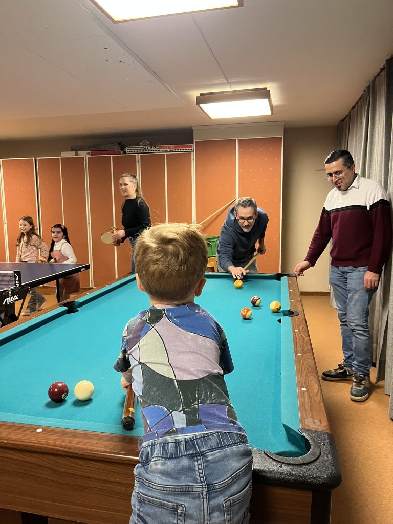 En ung pojke och tre vuxna spelar biljard, medan två flickor står i närheten i ett spelrum.