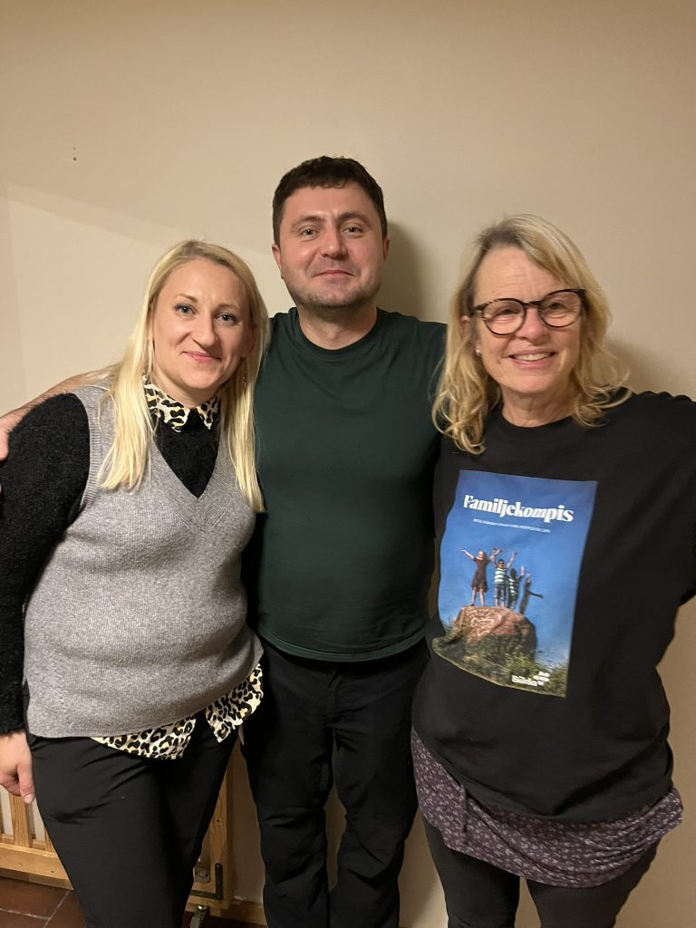 Tre vuxna står tillsammans inomhus och ler mot kameran med armarna runt varandra.