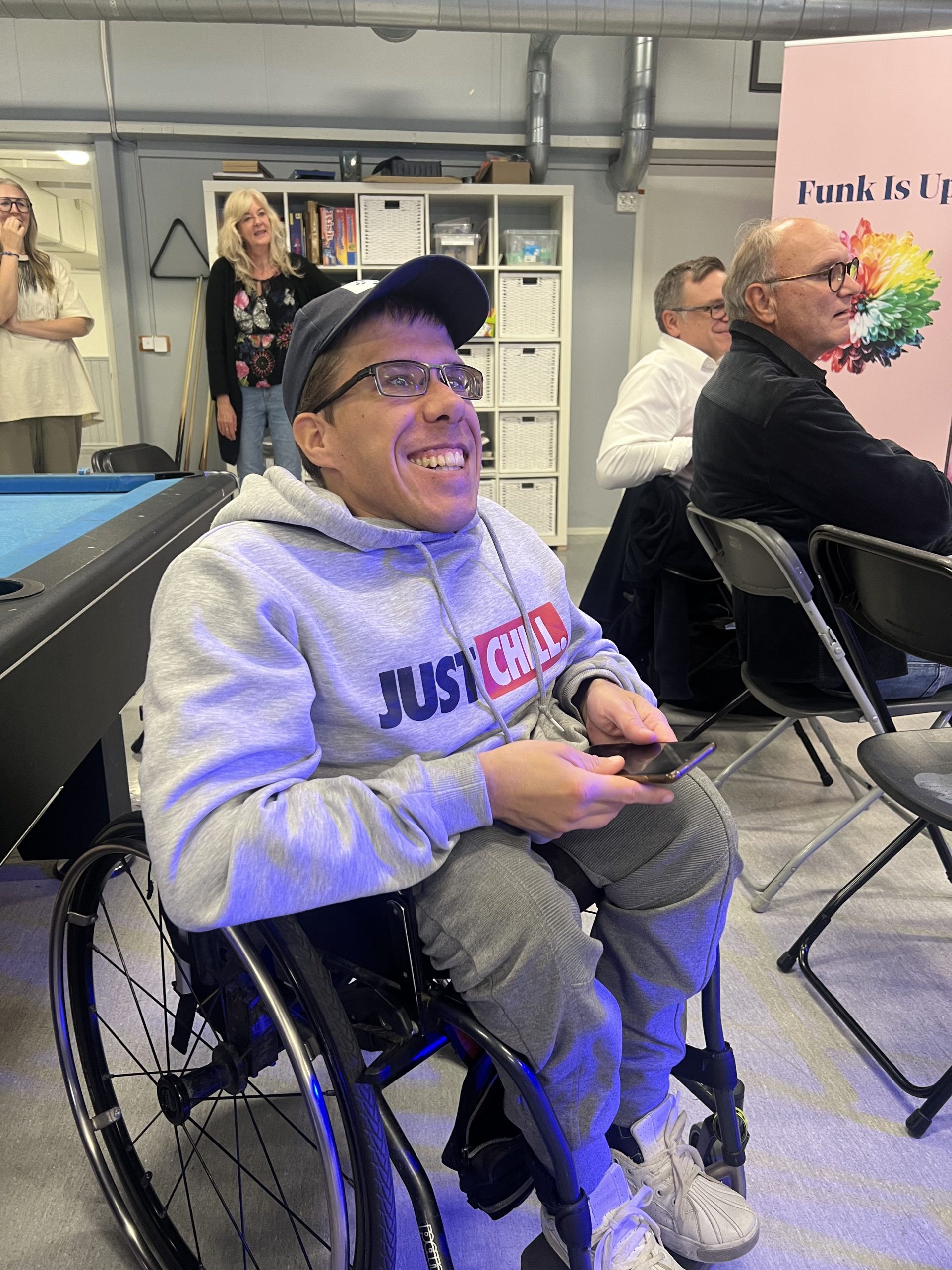 En leende man i rullstol med en "Just Chill"-hoodie sitter inomhus och håller en telefon i handen.