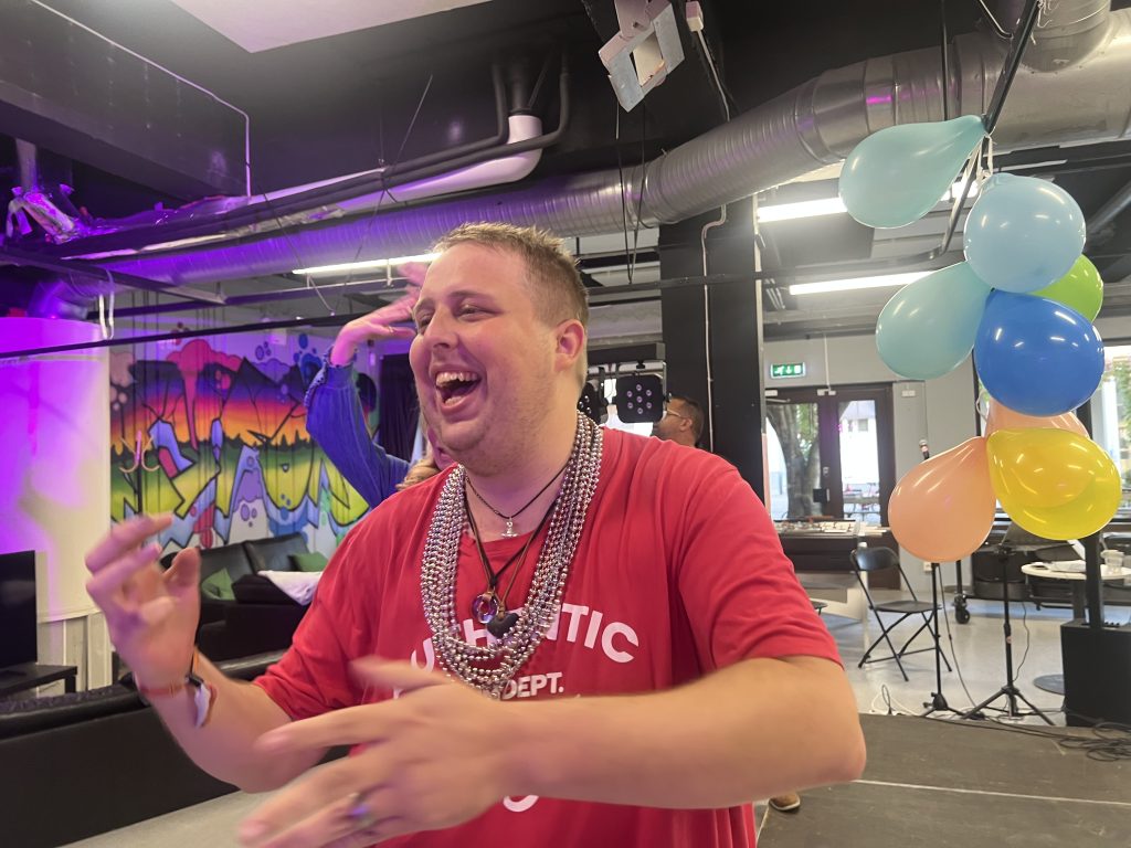 En man i röd skjorta ler och gestikulerar på en fest, med ballonger och färgglad graffiti i bakgrunden.