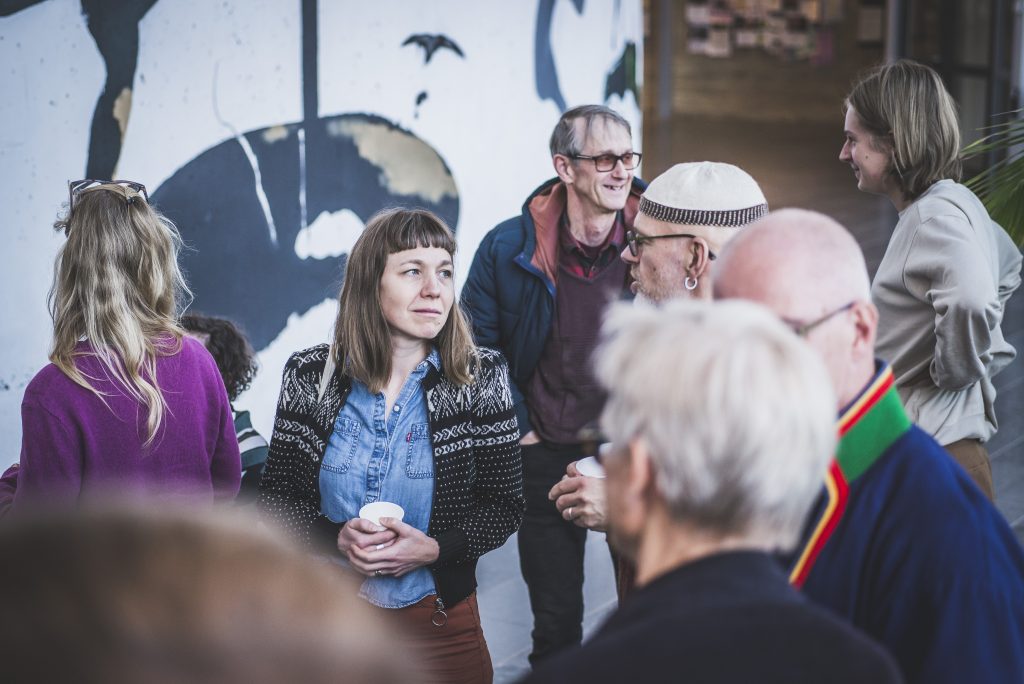En grupp människor står inomhus och pratar och ler under en avslappnad social sammankomst.