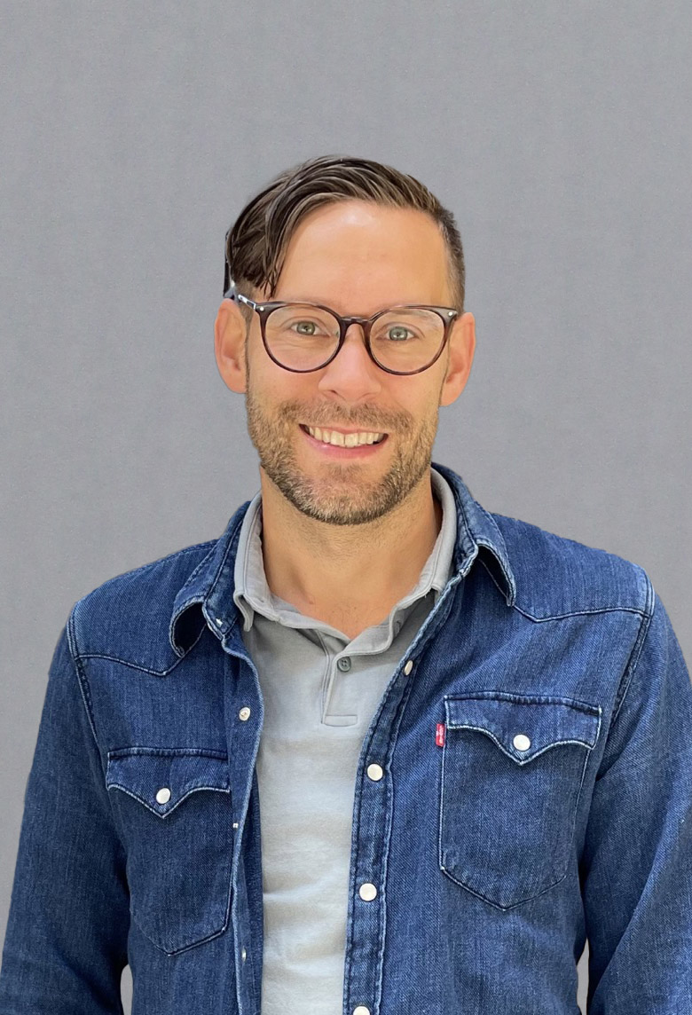 Leende man med glasögon, klädd i jeansskjorta över grå polo, står framför en grå bakgrund.