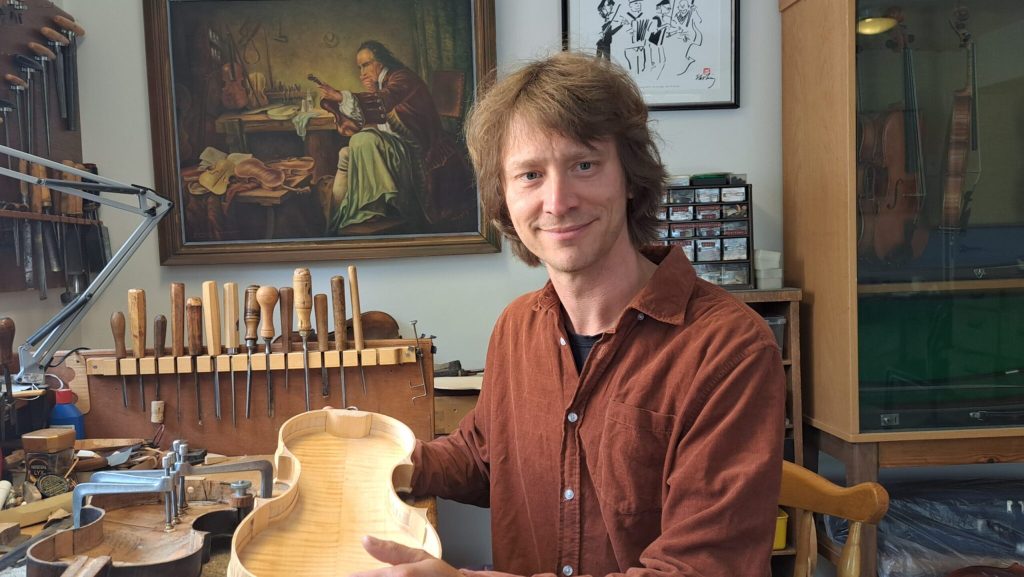 En man i en verkstad håller i en violinkropp, omgiven av verktyg och med målningar hängande på väggen bakom sig.