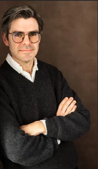 Man med glasögon och mörk tröja står med armarna i kors framför en brun bakgrund.