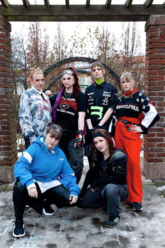 Sex unga människor i färgglada, tuffa streetwearkläder poserar självsäkert utomhus på en snöig stig mellan tegelpelare.