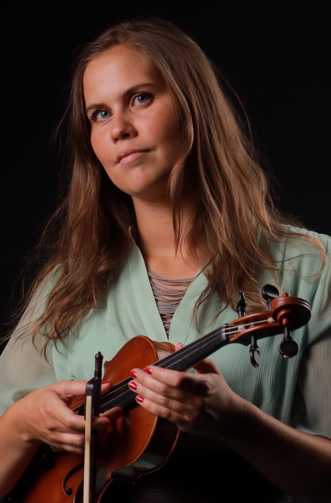 Kvinna med långt brunt hår håller en violin och stråke mot en mörk bakgrund, klädd i en ljusgrön blus.