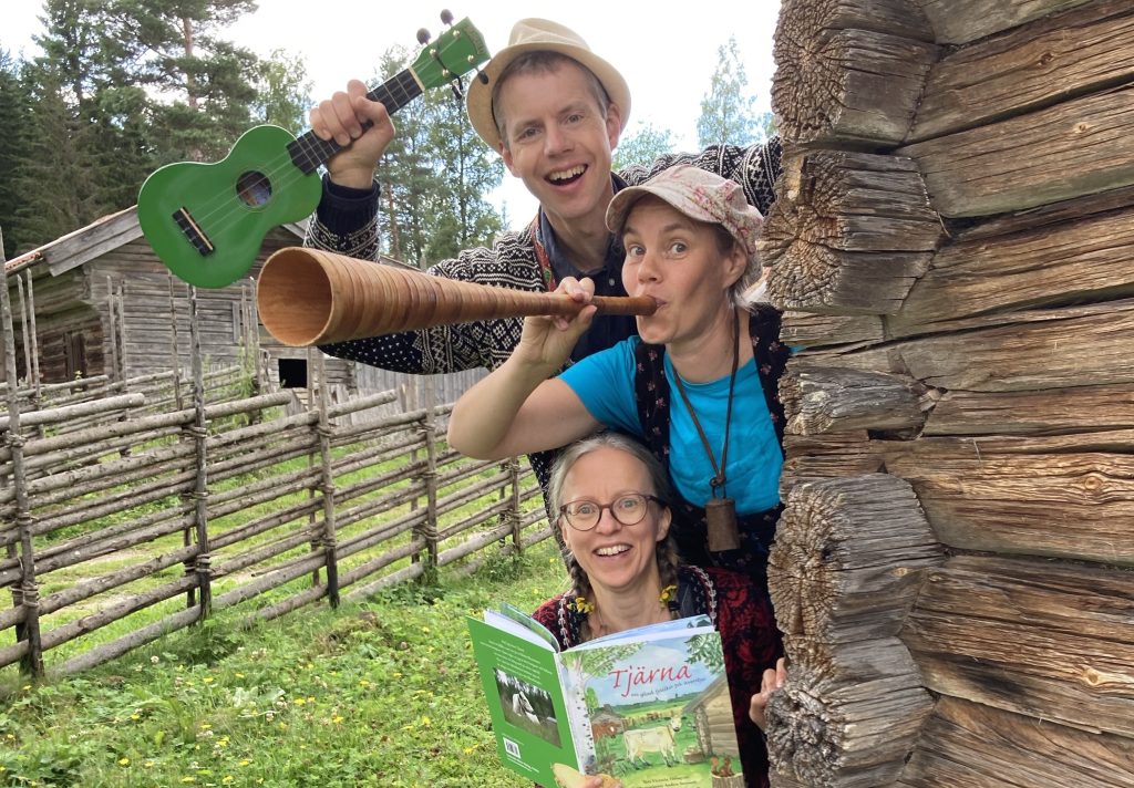 Tre glada människor vid en timmerstuga, en med ukulele, en som blåser i ett horn och en som håller i en öppen bok.