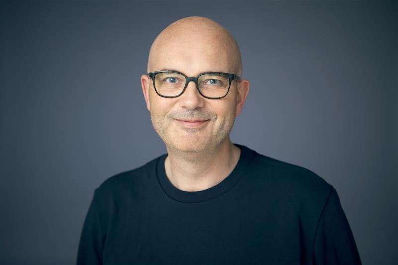 Leende skallig man med glasögon i svart skjorta mot en grå bakgrund.