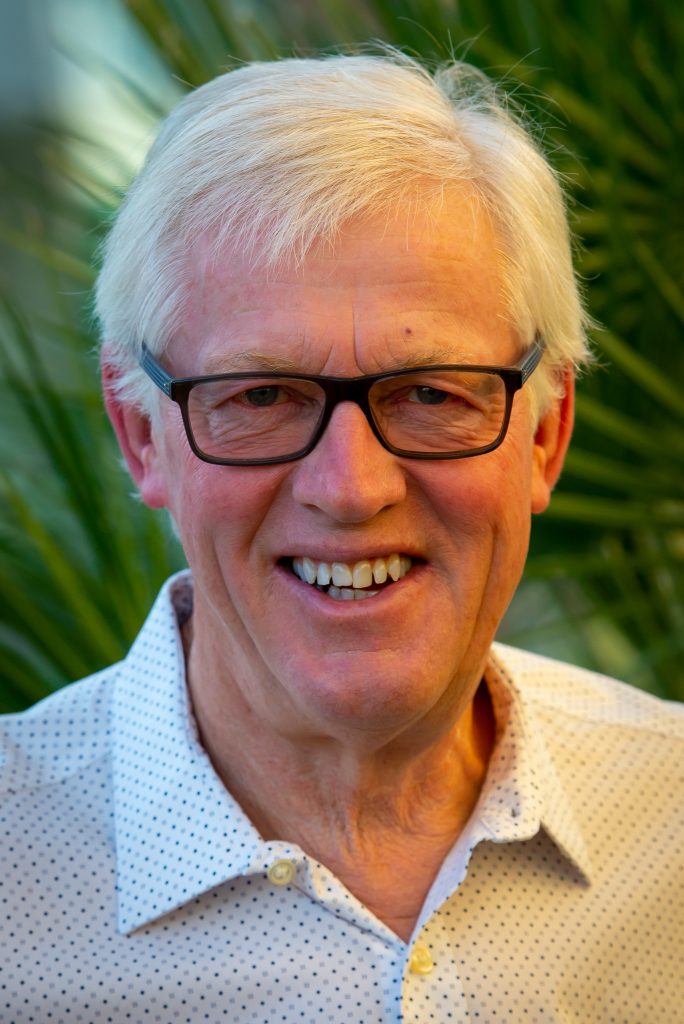 Leende äldre man med vitt hår och glasögon, klädd i ljus mönstrad skjorta, står utomhus bland grönska.