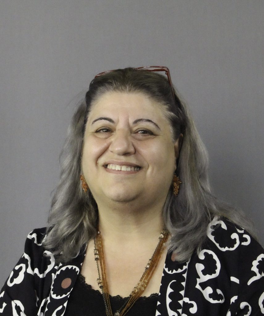 Leende kvinna med grått hår, glasögon på huvudet, mönstrad jacka och pärlhalsband mot en grå bakgrund.