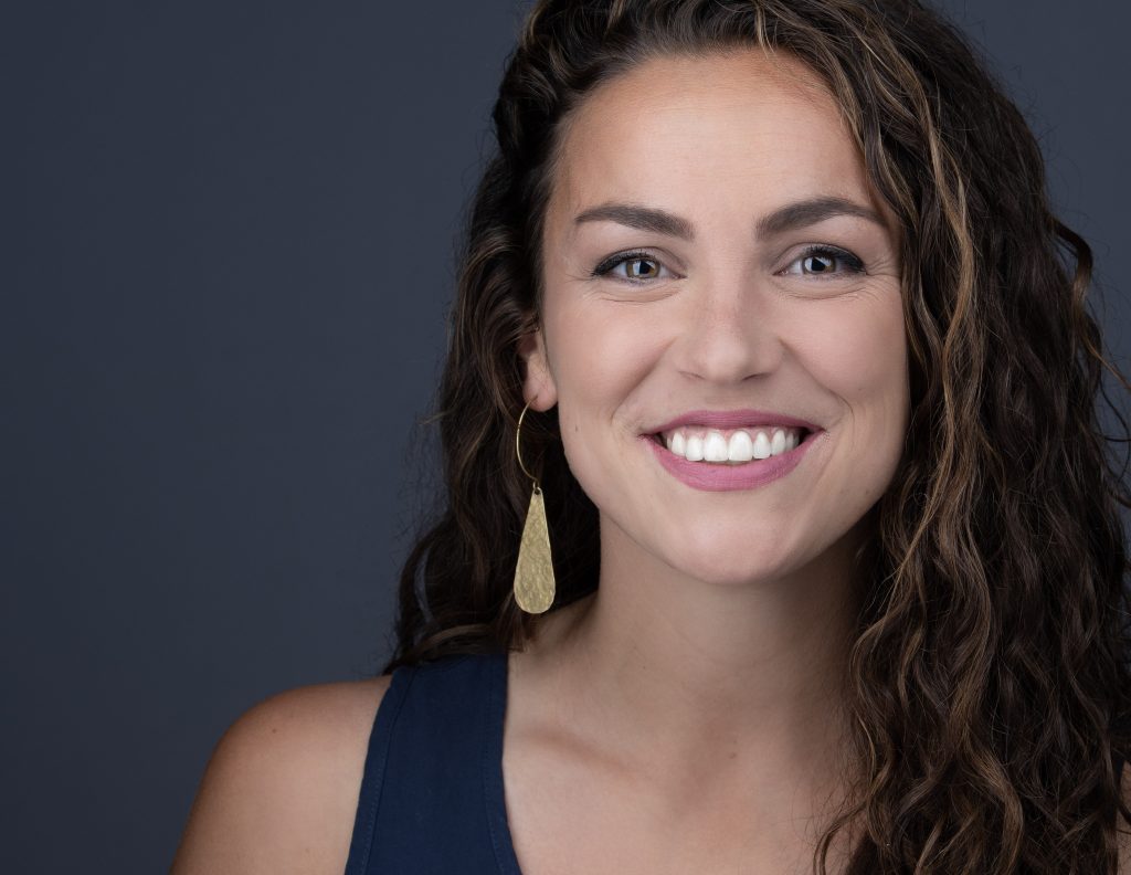 Leende kvinna med lockigt hår, bär guldörhänge och marinblå topp, mot mörk bakgrund.