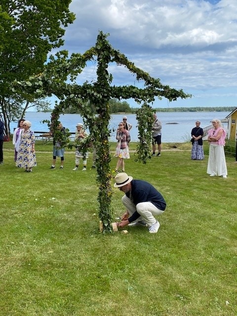 Människor samlas utomhus medan en man dekorerar en lövklädd majstång på en gräsmatta nära vatten under en molnig himmel.