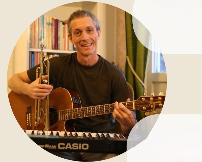 En leende man spelar gitarr, håller i en trumpet, med en keyboard framför sig och bokhyllor i bakgrunden.