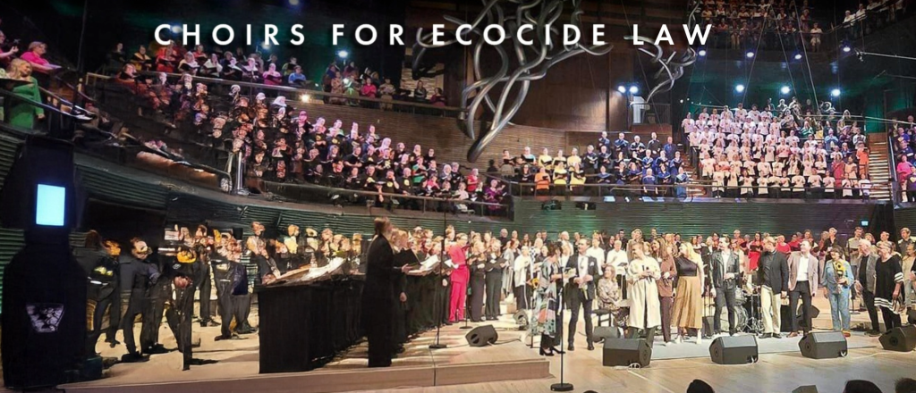En stor kör uppträder på scen under texten "Choirs for Ecocide Law" i ett modernt konserthus.