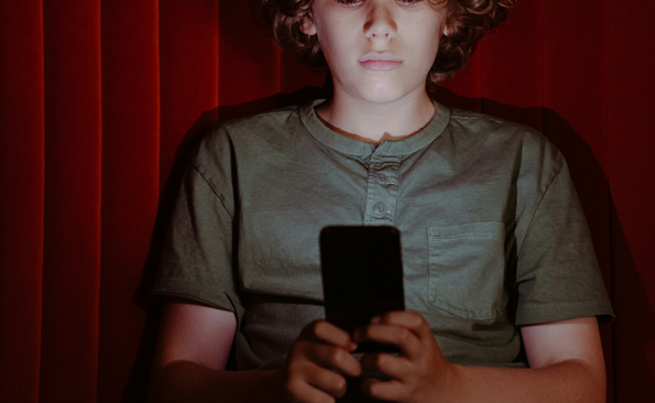 En pojke med lockigt hår tittar på en smartphone, upplyst av dess ljus, mot en röd bakgrund.