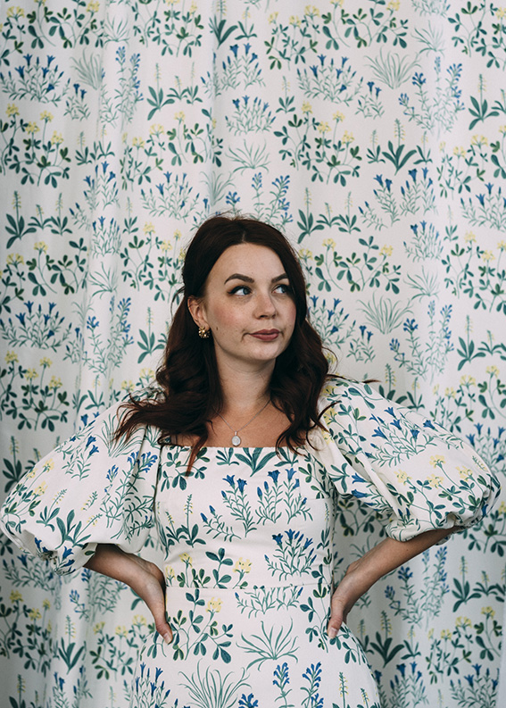 Kvinna i blommig klänning står framför en matchande blommig bakgrund med händerna på höfterna och tittar åt sidan.