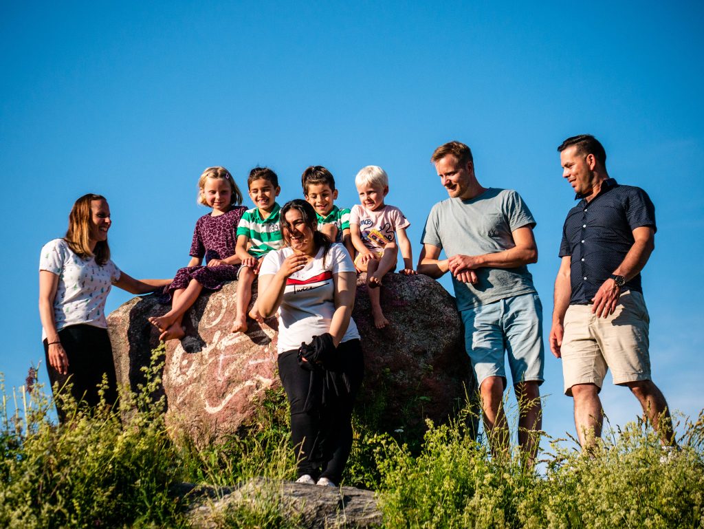 Sex vuxna och fyra barn ler och sitter på en stor sten utomhus under en klarblå himmel.