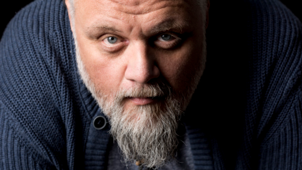 En man med grått skägg och blå tröja tittar uppmärksamt in i kameran mot en mörk bakgrund.